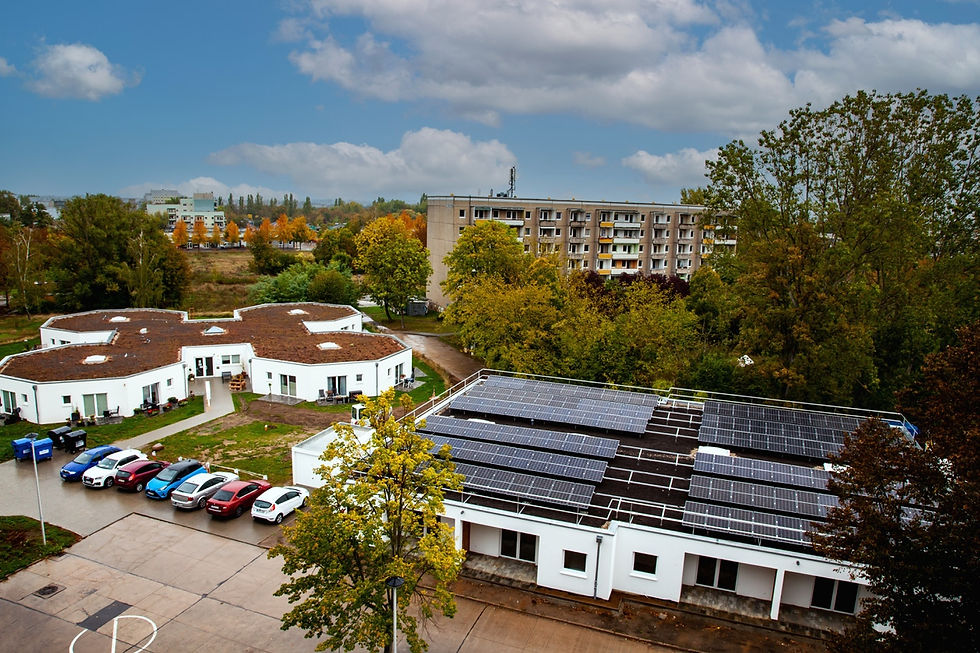Rooftop solar panels on white buildings, surrounded by autumn trees. Several parked cars and an apartment block in the background. Cloudy sky.