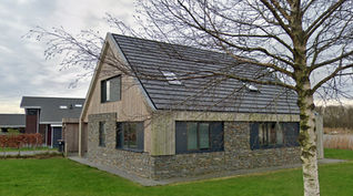 Modern house with a combination of stone and wood facade, featuring a steeply pitched dark roof with skylights. Large windows provide natural light, and the house is surrounded by a green lawn with a leafless tree in the foreground. The background includes other contemporary homes and an overcast sky.