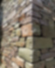 A Welsh sandstone wall. The stone is a mixture of dark reds, greens, browns and oranges. The wall is shy-bedded, giving the appearance of a mortarless dry-stone wall.