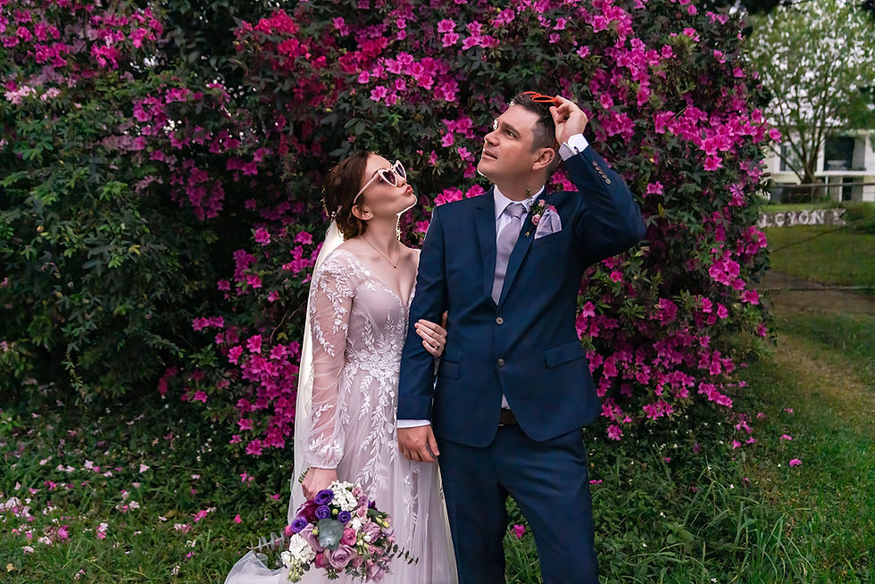 Couple in formal attire with sunglasses, posing by colourful pink flowers. Bride holding bouquet, both looking upwards, cheerful mood.