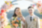 Bride and groom smiling under a colorful floral arch, she holds a vibrant bouquet. Bright outdoor setting at the Lakehouse Folk Sunshine coast with a serene backdrop.