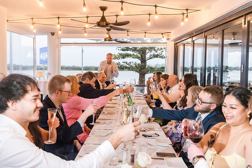 People toast at a long dining table in a well-lit balcony with large windows and a view of trees and water, creating a joyful atmosphere.