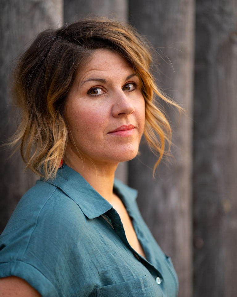 Portrait headshot of Nicola Elson in a teal shirt against a soft, blurred wooden background