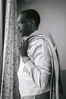 Groom with chronograph watch holds coat jacket over shoulder while looking out the window of hotel room.