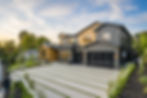 Modern custom home with black and wood exterior, large windows, glass garage doors, and landscaped driveway at sunset.