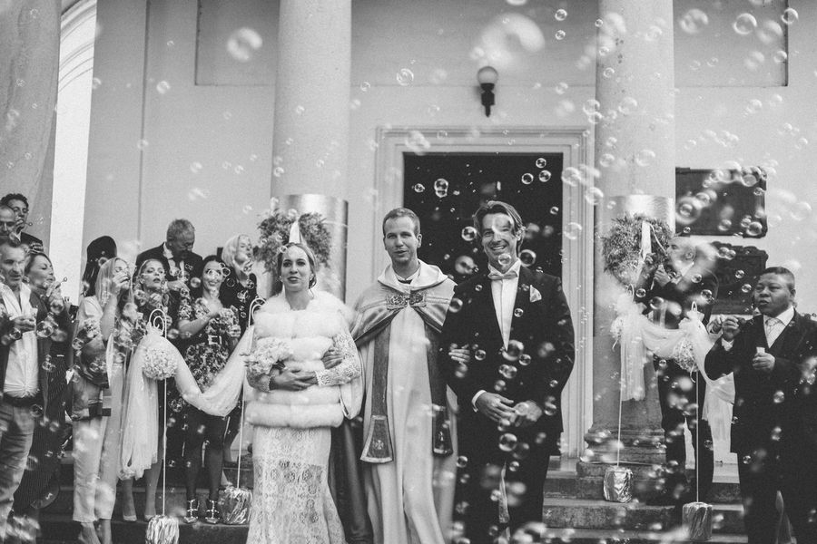 While uncountable bubbles fly in the air, priest leads happy wedding couple out of church after being united in matrimony.