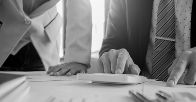 A imagem é uma fotografia em preto e branco que captura um ambiente de trabalho corporativo e analítico. Em primeiro plano, o foco está nas mãos de dois indivíduos vestidos com trajes profissionais (ternos e blazers).
