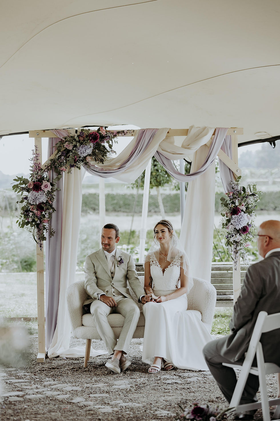 Bruidspaar bij ceremonie op bankje voor backdrop met stof en bloemen