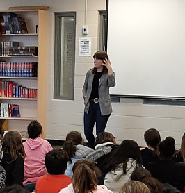 Canadian children's book author C.A. Klug talks to a school. Classrooms of children listen and ask questions.