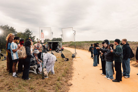 Film crew and cast members standing on a dirt road during an outdoor video production with large white diffusion screens and equipment setup.