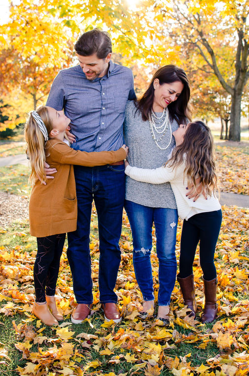 A family of four stands together in a vibrant autumn setting, surrounded by colorful leaves, sharing warm embraces and smiles.