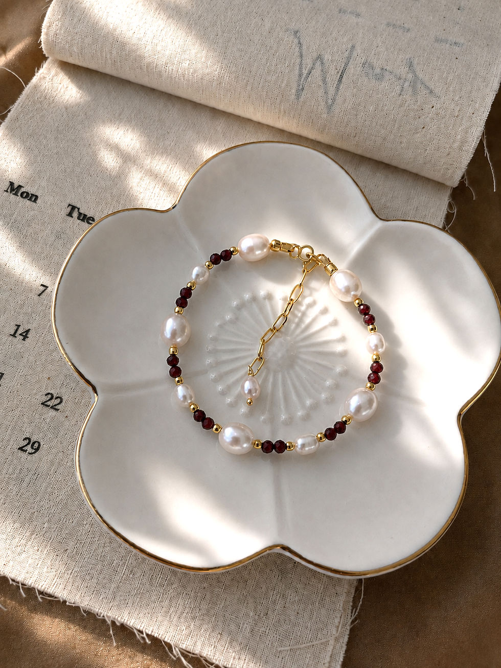Cranberry garnet and freshwater pearl bracelet on a gold-rimmed ceramic dish, with a calendar page in the background