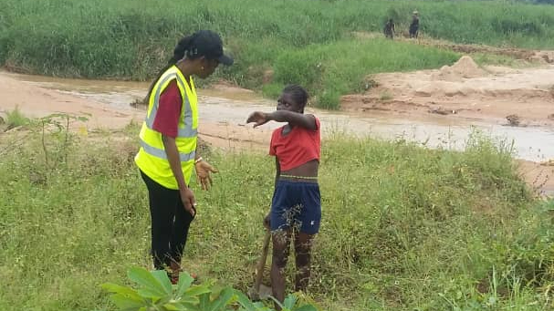 AIG DRC representative in a reflective vest engaging in conversation with a young girl near farmland.