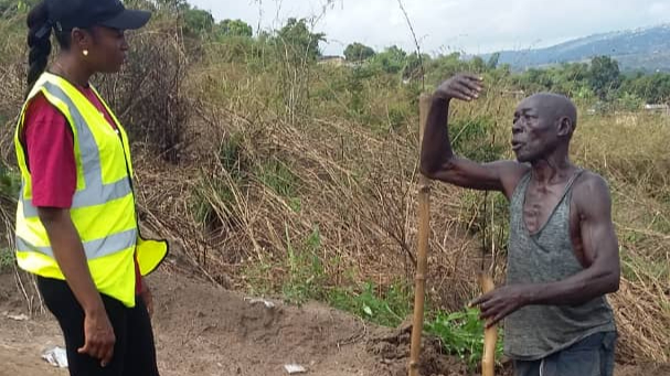 AIG DRC staff member in a reflective vest speaking with an elderly man holding a tool during a field visit.