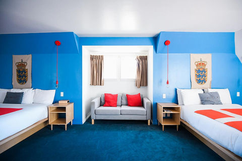 Hotel room with two custom bed-frames and nightstands. Room is vibrant blue with velvety carpet and red and white decor.