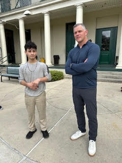 a man in a blue longsleeve shirt and blue pants stands with his arms crossed next to a child
