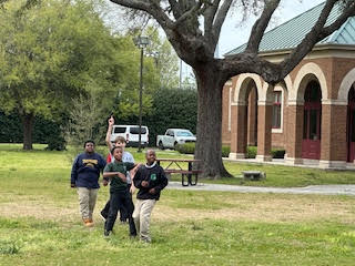 three children stand in a grassy field. There is a tree and a red brick building in the background