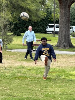 a child kicking a ball while in a grassy field. a man in a light blue shirt is walking across the background