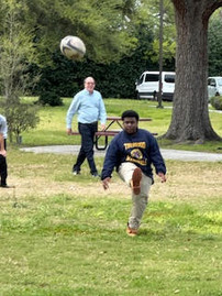 a child kicking a ball while in a grassy field. a man in a light blue shirt is walking across the background