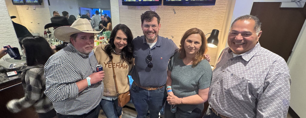 three men and two women pose in a small room. One man has on a cowboy hot. There are two screens in the background