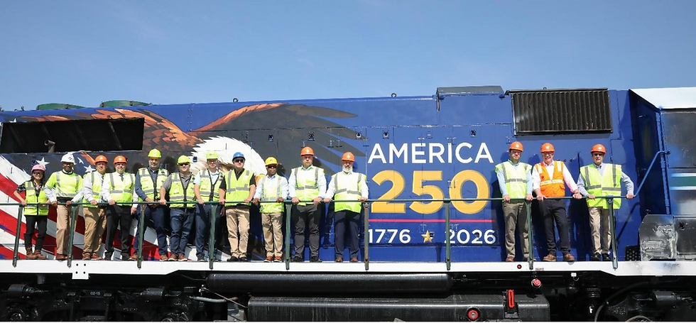 Railroad workers in safety vests and hard hats pose on a blue locomotive with America 250, 1776–2026, under a clear sky.