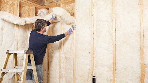 Man in blue shirt installing insulation in a wooden wall while standing on a ladder. Bright, well-lit setting, focused work atmosphere.