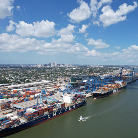 Cargo ships labeled CMA CGM and MSC docked at a busy port with colorful containers under a partly cloudy sky. City skyline visible in background.
