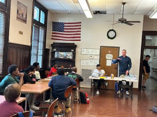 a man in the front of a classroom speaks to children at tables around the room