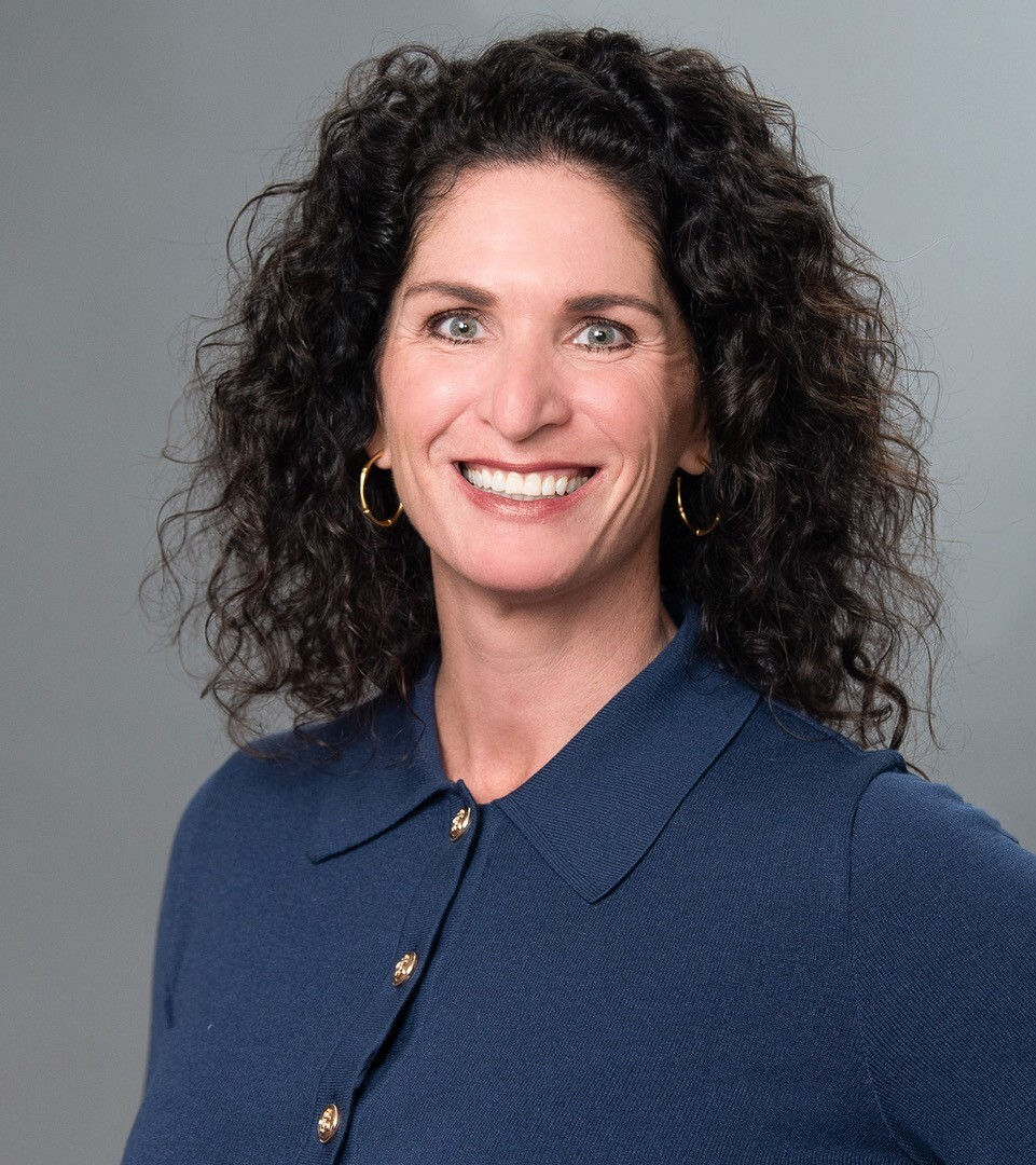 Smiling woman with curly hair in a blue collared shirt against a plain gray background. She wears hoop earrings and exudes confidence.