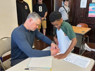 a man sits at a table and signs an autograph on a child's shirt