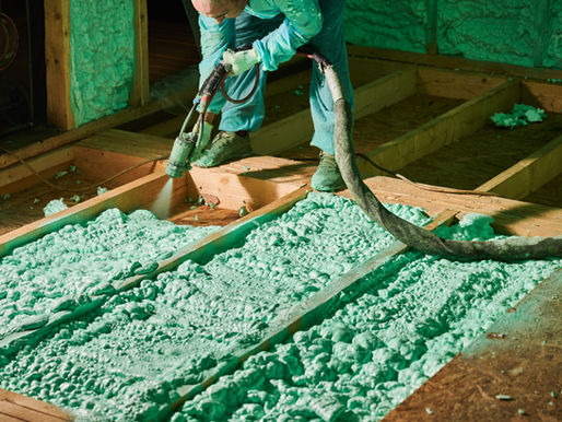 Worker sprays green foam insulation between floor joists in a wooden structure. The setting is dimly lit, with a focused, industrial atmosphere.