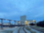 Train yard at dusk showing rail tracks curving towards a lit industrial building. Tanker trucks labeled "Suttons" and train cars visible.