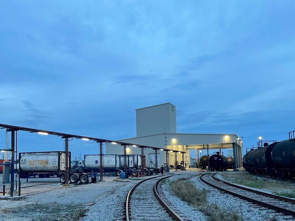 Train yard at dusk showing rail tracks curving towards a lit industrial building. Tanker trucks labeled "Suttons" and train cars visible.