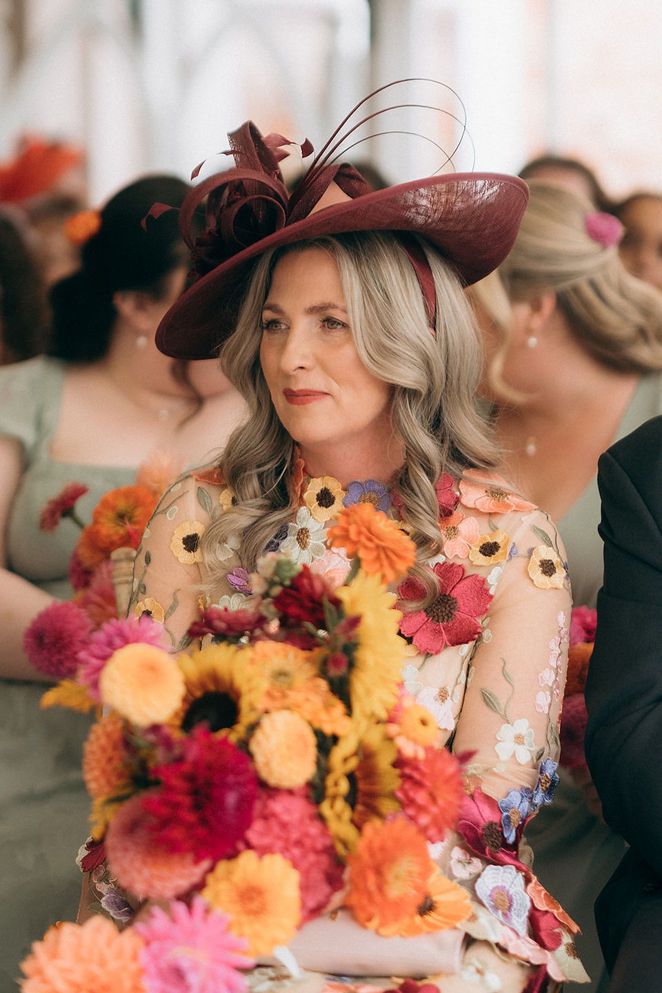 The mother of the bride holding the bride's hand-tied bouquet, featuring a vibrant mix of textured dahlias, bright zinnias, and bold yellow sunflowers designed by Jacqui O. The arrangement is full of warm pink, orange, and yellow summer tones.