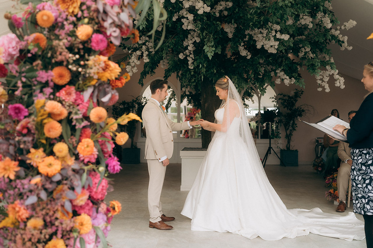 A bride and groom exchanging ring at Combermere Abbey Glass House with colourful flowers by Jacqui O 