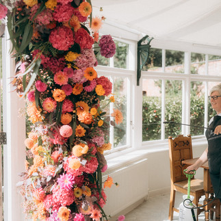 A wedding ceremony at Combermere Abbey featuring a "broken arch" floral installation by Jacqui O. The arrangement is composed of dahlias, zinnias, and hydrangeas in vibrant shades of pink and orange with yellow accents.