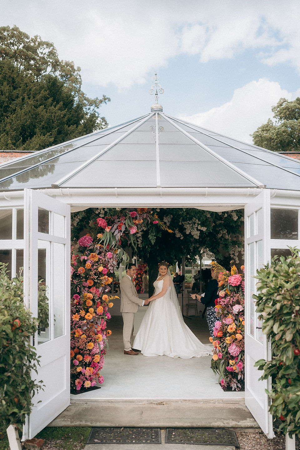 A joyful bride and groom celebrate their marriage at Combermere Abbey. They stand before a striking asymmetrical floral arch of dahlias and zinnias in a warm sunset palette of pink, orange, and yellow.