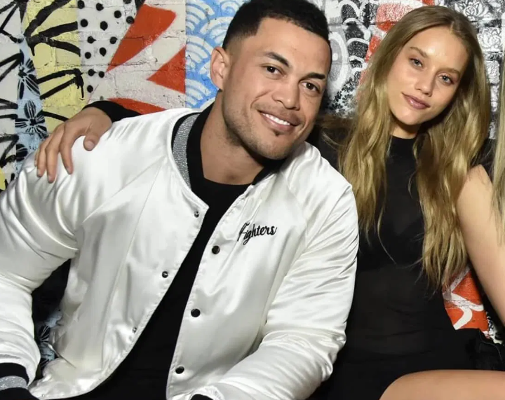 Giancarlo Stanton and Chase Carter sit closely, smiling, against a colorful graffiti wall. The man wears a white jacket with "Fighters" text. Casual atmosphere.