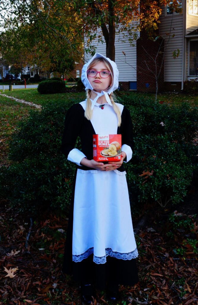 A girl with glasses dressed in a pilgrim costume looking to the left. She holds a Tofurkey box.