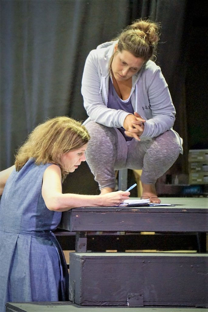Sally and Pierrette Swan during Tempest rehearsals. They are on a platform looking at a script, the background is a black curtain.