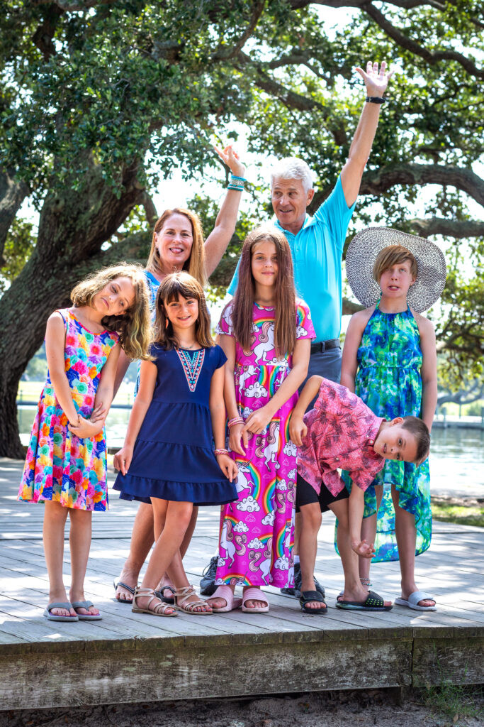 A man and woman with arms their left arms in the air. Four gilrs stand. On boy bends over to the left side facing the camera. The last child is a girl in a white hat.