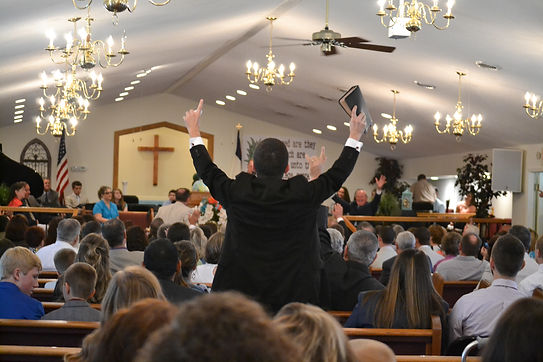 Man with arms raised, holding Bible, speaks to Life Baptist Church congregation.