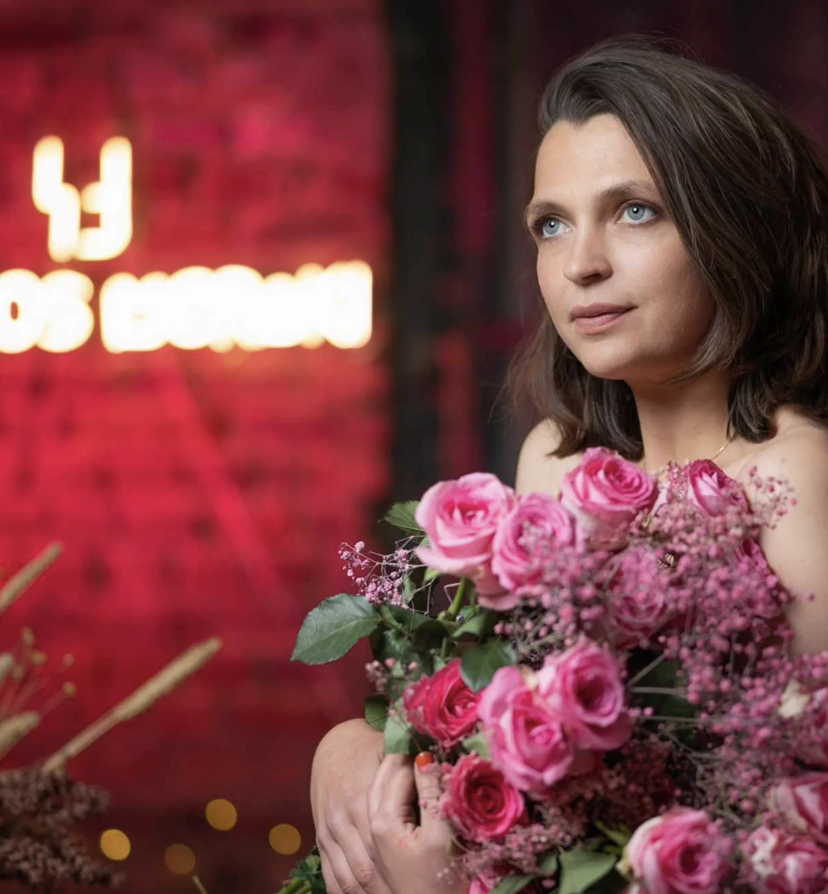 Portrait de Constance Montaigne tenant un grand bouquet de roses roses, posant devant un mur en briques illuminé de rose avec le néon "Le Clos Meraki" en arrière-plan. Photo prise lors de l’afterwork Octobre Rose 2023 dans une ambiance douce, intime et engagée.