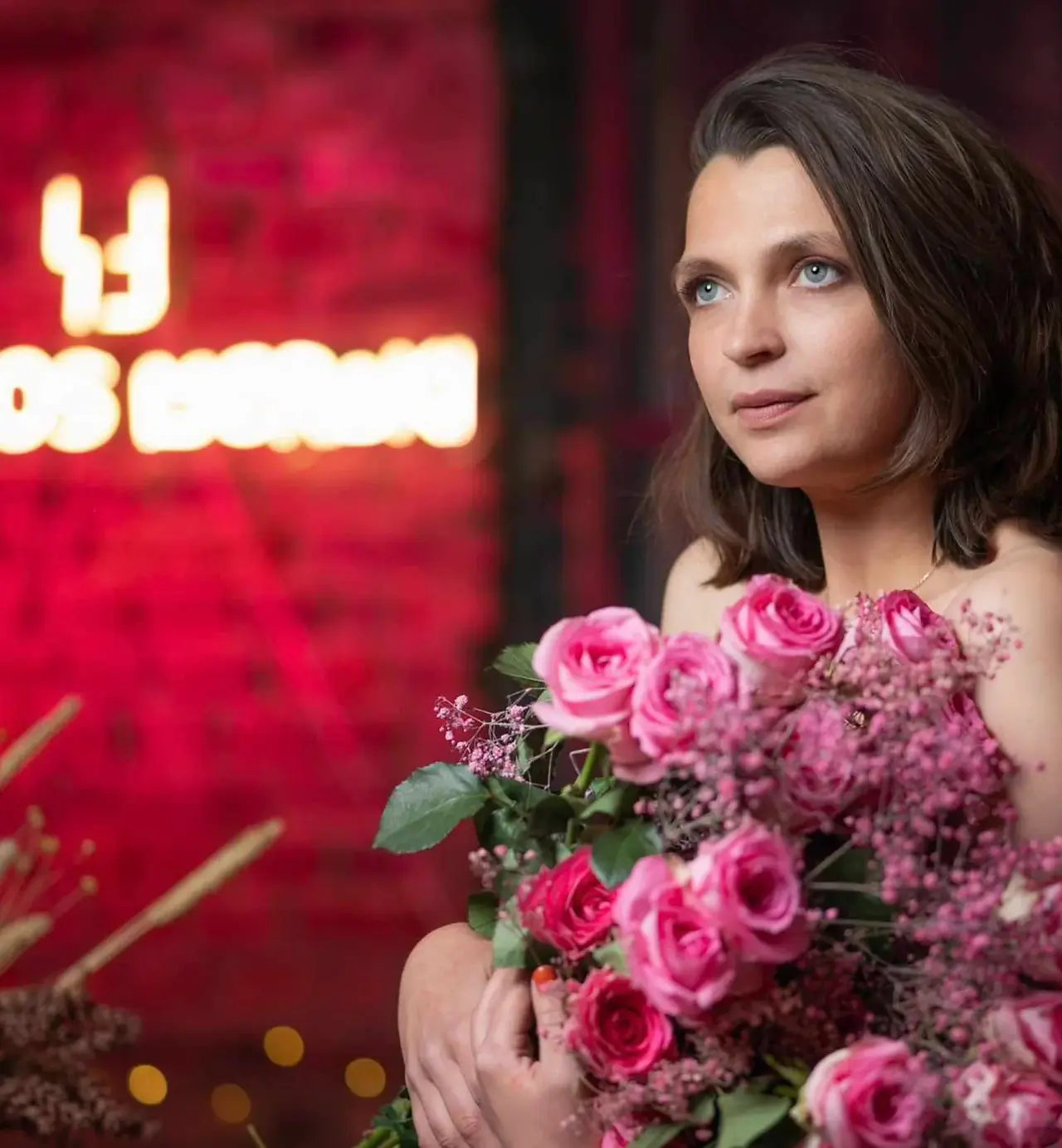 Portrait de Constance Montaigne tenant un grand bouquet de roses roses, posant devant un mur en briques illuminé de rose avec le néon "Le Clos Meraki" en arrière-plan. Photo prise lors de l’afterwork Octobre Rose 2023 dans une ambiance douce, intime et engagée.