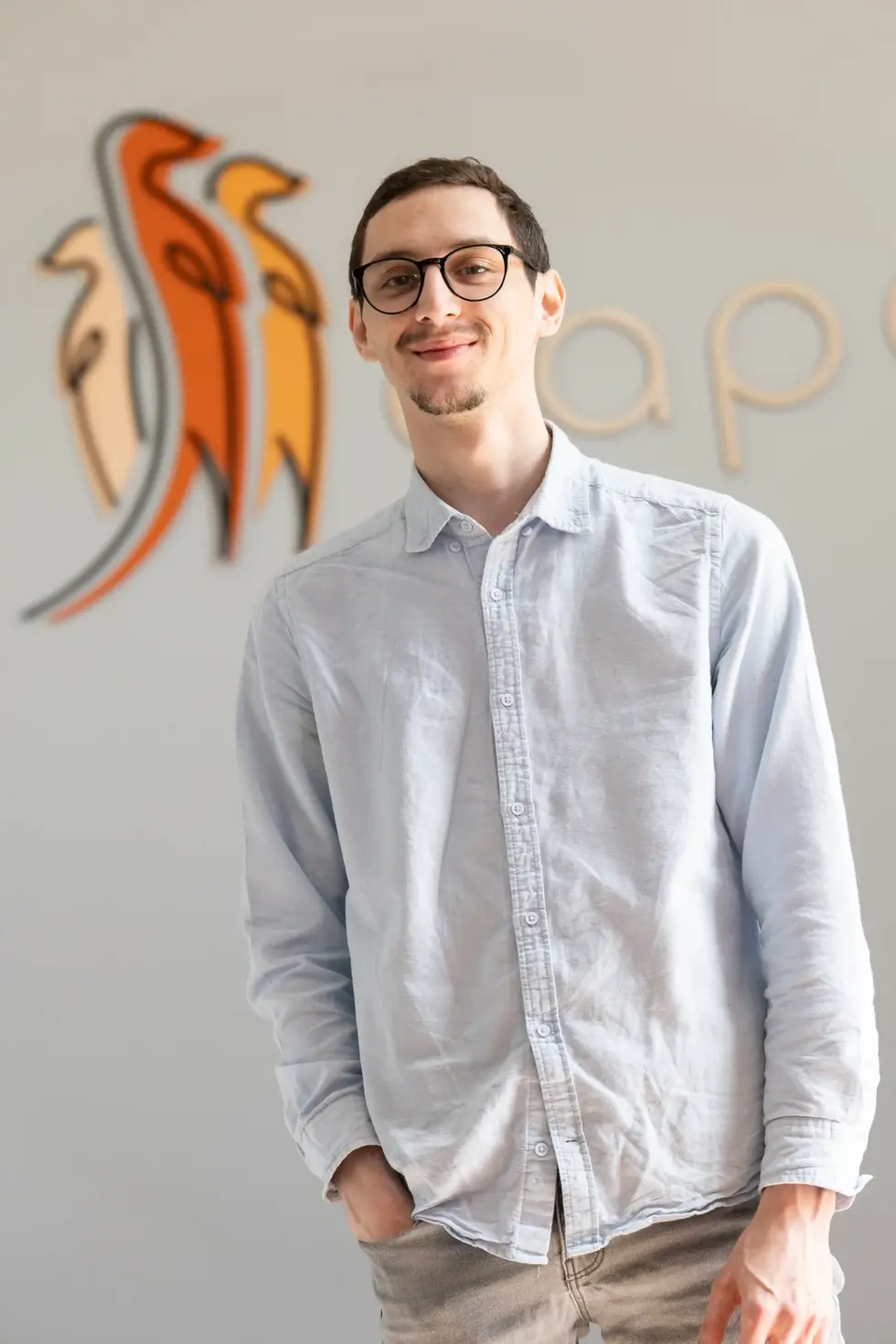 Portrait d’un collaborateur de Capeoni souriant, debout devant le mur d'accueil avec le logo de l'entreprise en arrière-plan.