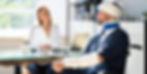 A man with head and arm injuries, wearing a neck brace and arm cast, sits in a wheelchair at a desk across from a woman, possibly discussing a personal injury case