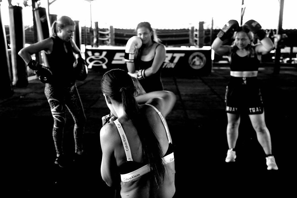 Черно-бяла снимка показва четири жени във фитнес зала, три от които носят боксови ръкавици, което показва тренировка или сесия по бойни изкуства като муай тай или кикбокс.