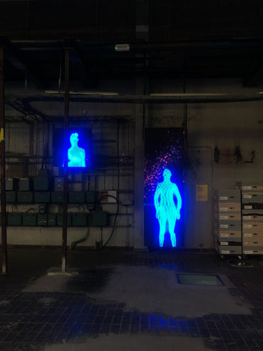 Two blue glowing digital human figures on separate vertical screens in an industrial space, one full body and one partial figure