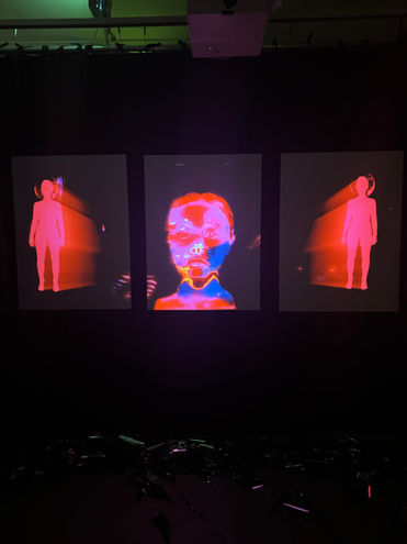 Three vertical screens showing a digital face and human figures in red tones in a dark exhibition space