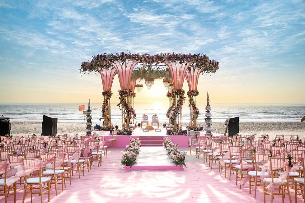 **Alt text:**
A stunning beachside wedding setup featuring a grand pink mandap adorned with cascading flowers and chandeliers, set against a serene ocean backdrop at sunset. The aisle is lined with floral arrangements and gold chairs draped in pink fabric, creating a dreamy and elegant ambiance.